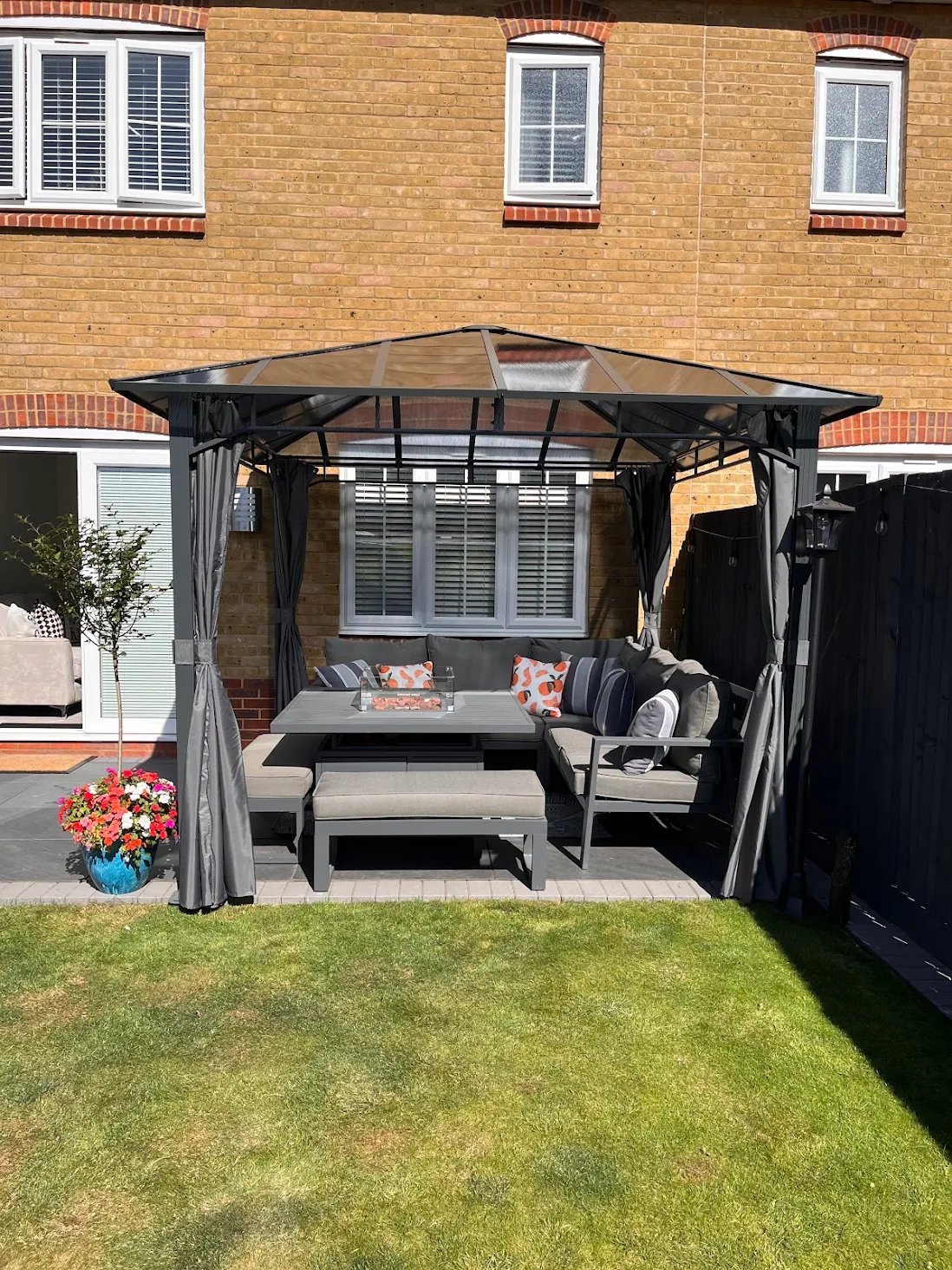 Black garden gazebo with shade canopy over seating area and table on green lawn in front of brick house